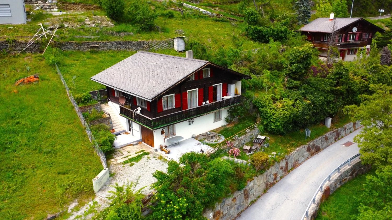 Two-story chalet-style house with red wooden exterior, surrounded by lush greenery and a stone wall. The house has a balcony and is situated on a hillside with a scenic view.