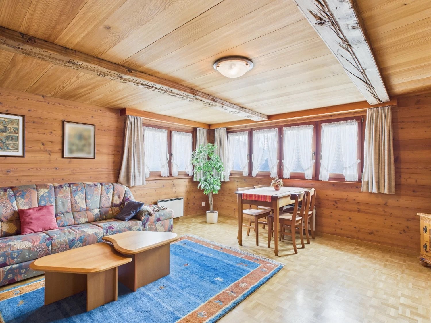 Cozy wooden cabin-style living room with large windows, floral patterned sofa, wooden dining table and chairs, and a fireplace