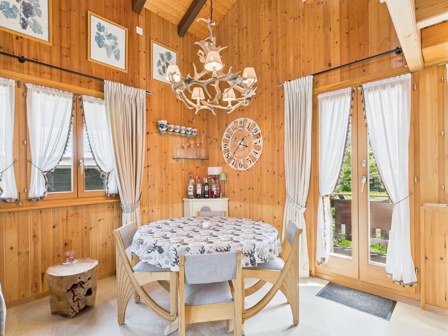Cozy wooden cabin-style dining room with a large antler chandelier, a round dining table with patterned tablecloth, and chairs. The room has wooden walls and ceiling, large windows, and a fireplace.