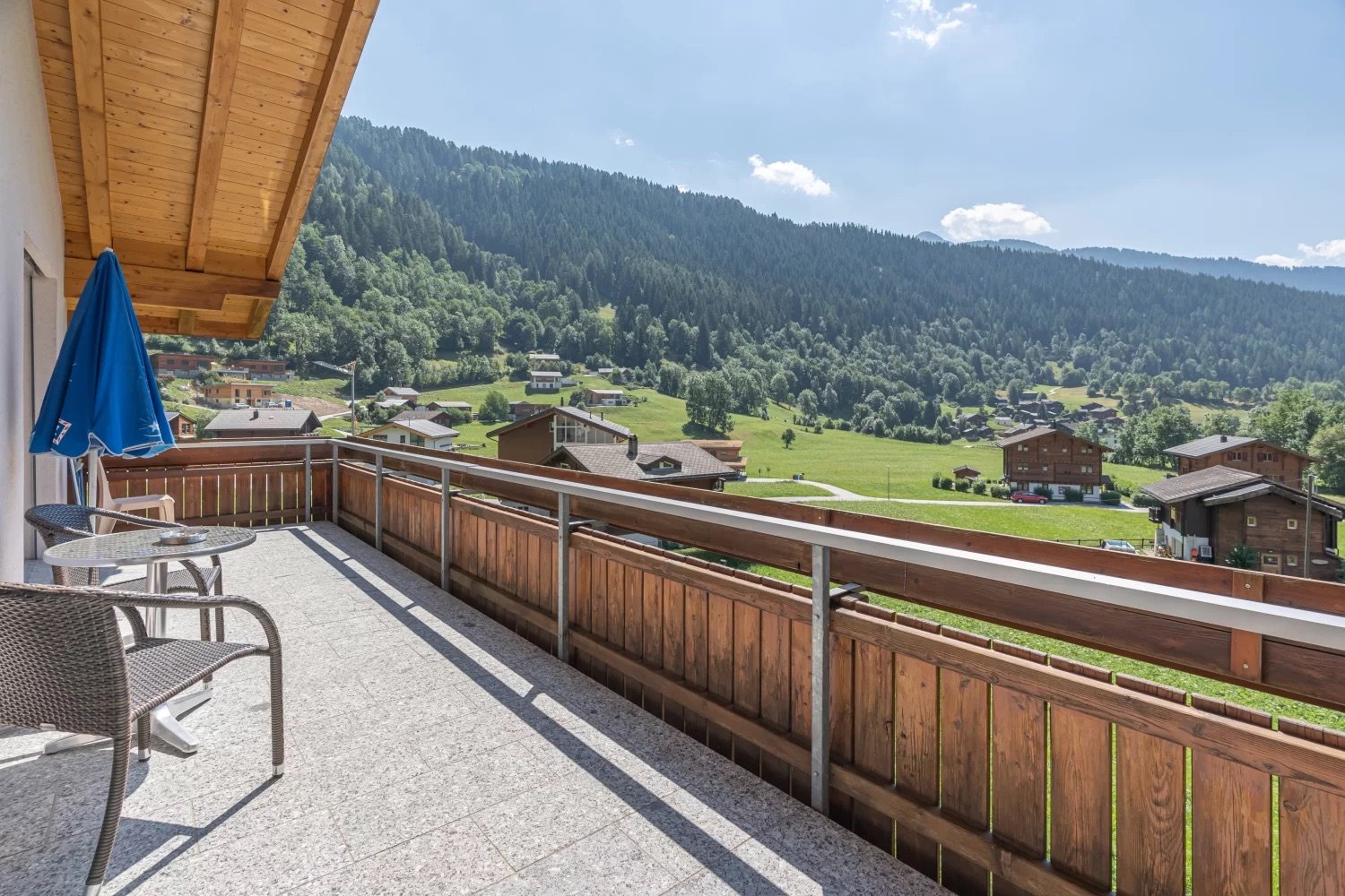 Wooden balcony with a view of a mountainous, forested landscape. The balcony has a table and chairs, and overlooks a village nestled in the valley below.