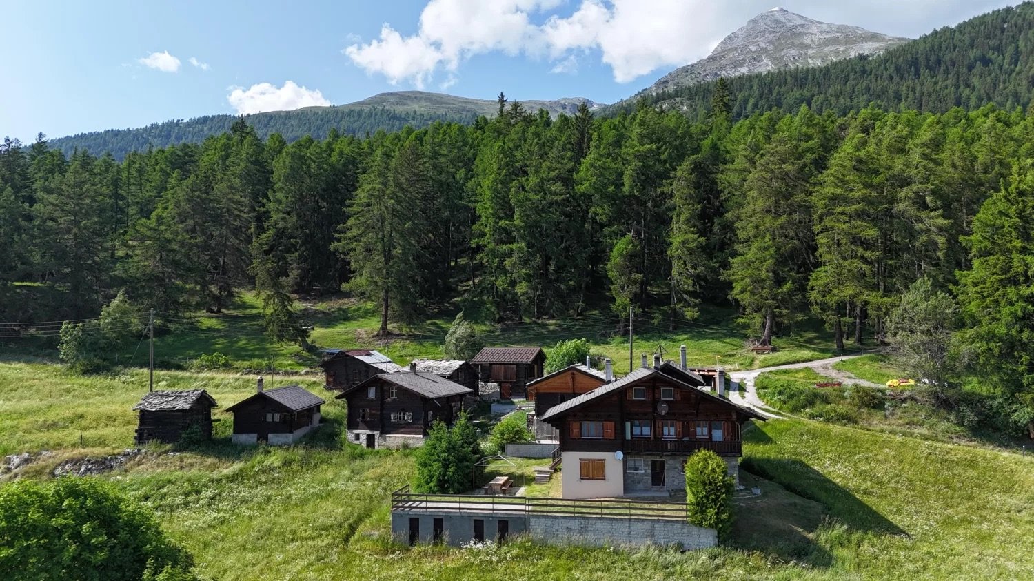 The image shows a picturesque mountain village nestled in a lush, green valley surrounded by towering mountains and dense forests. The village consists of several traditional wooden chalets and houses, some with balconies or terraces. The buildings are si