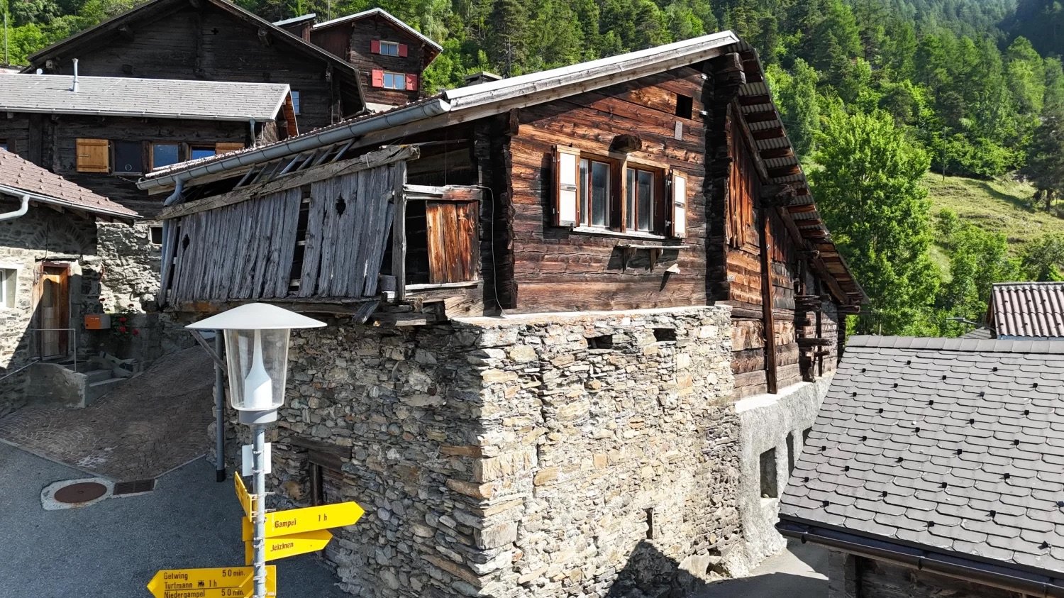 Traditional alpine-style wooden cabin with stone foundation, surrounded by lush green forest. The cabin has a balcony or terrace, and there are directional signs indicating nearby destinations.