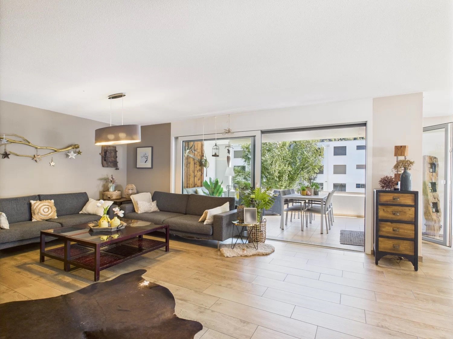 Spacious open-plan living room with large windows overlooking a balcony or terrace, grey sectional sofa, wooden coffee table, and a dining area with a table and chairs. The room has a modern and cozy decor with wooden accents and decorative elements.