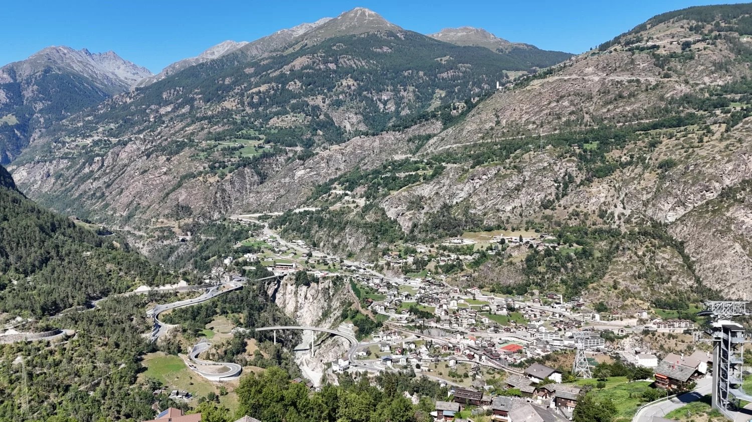 Mountain valley with a town below, winding roads, bridges, surrounding trees, mountains in the distance, clear sky.