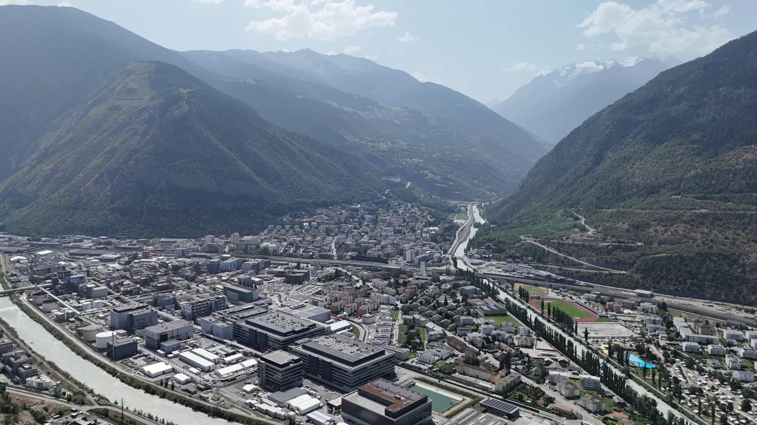 Aerial view of a city with mountains in the background, large buildings, wide river, and trees and plants lining the streets.
