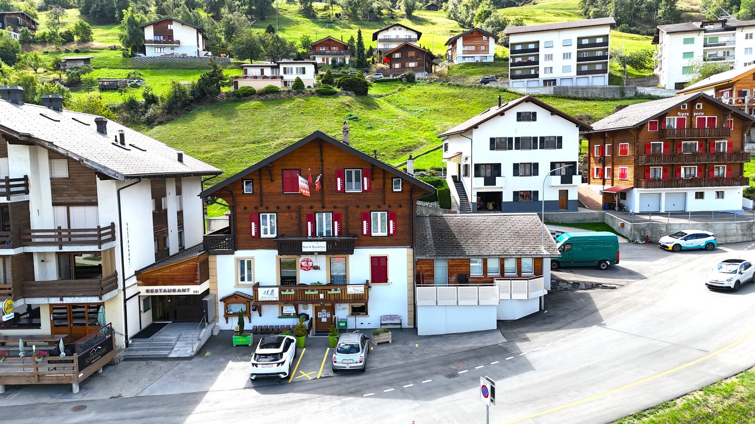 Mountain village complex with multiple buildings, wooden facades, and red shutters. One building has a restaurant sign. The complex is surrounded by a green hillside. Cars parked in front of the buildings.