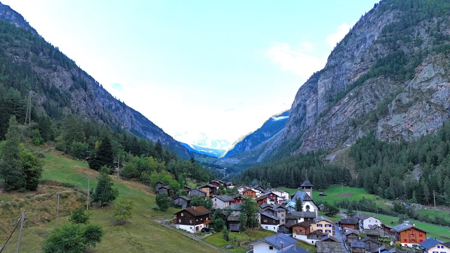 cluster of houses, green trees, rocky mountains, church, grassy fields