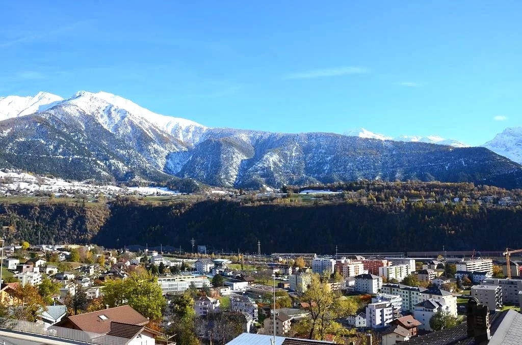 View of mountains with snow in background, surrounded by a town with various buildings, trees and greenery
