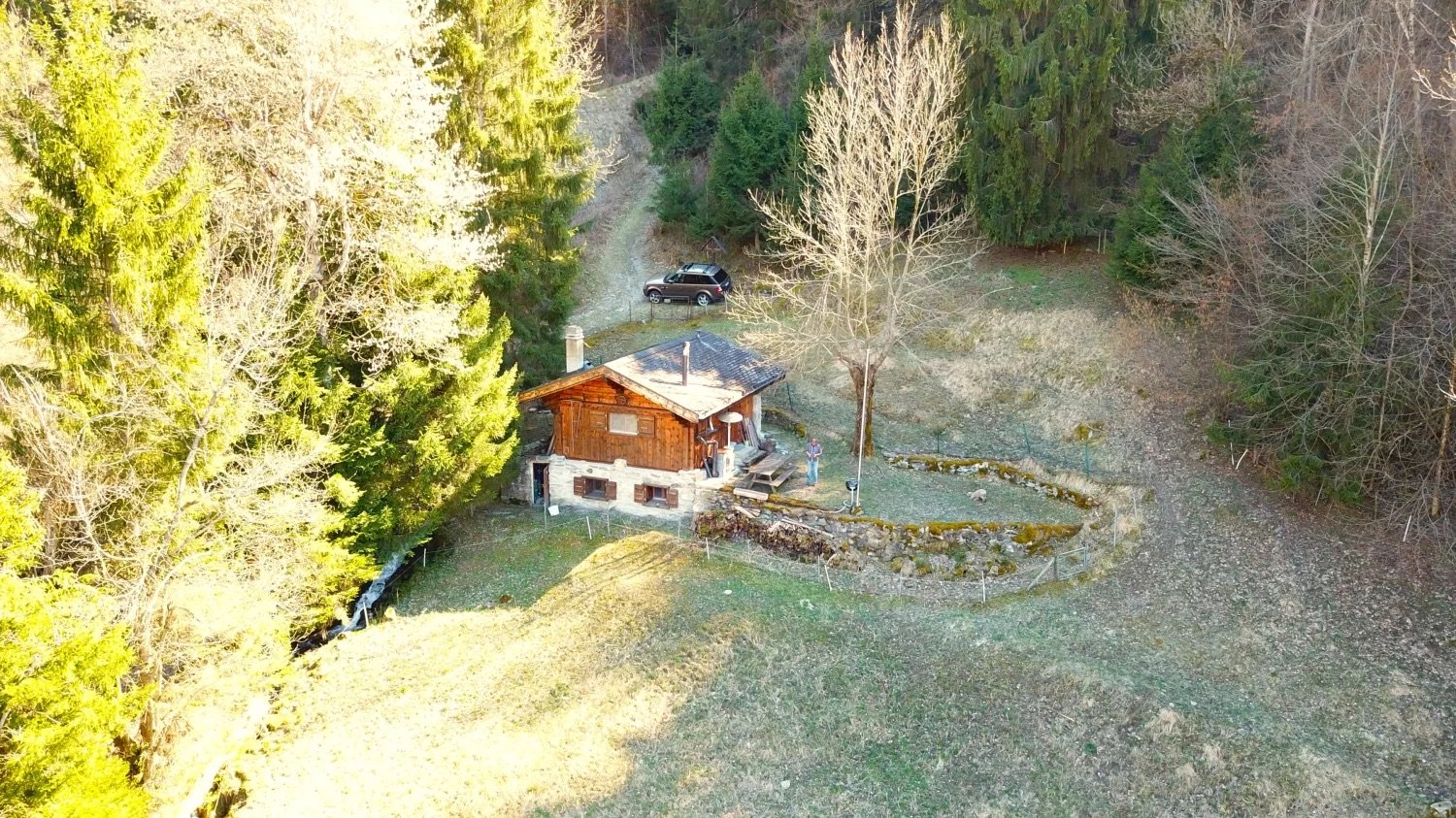 A small wooden house surrounded by trees, a car parked in front, stone walls and a wooden fence.