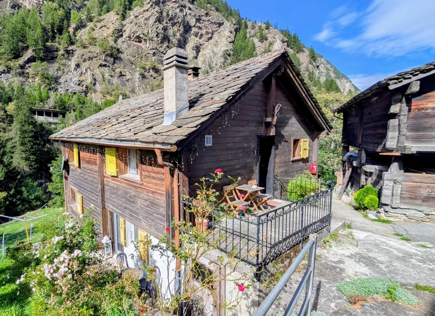 Small wooden chalet with a stone roof, yellow shutters, a chimney, a small balcony with a table and chairs, surrounded by plants and flowers, with a scenic mountain view