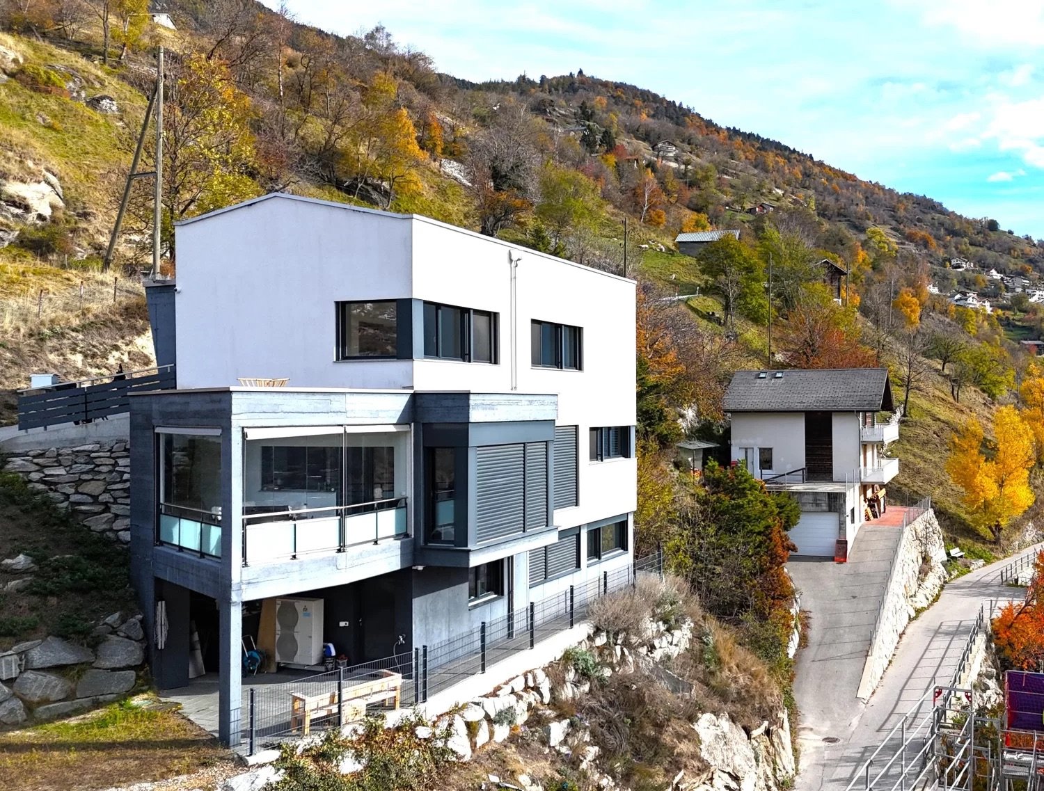 2 story modern house, raised ground floor, white exterior, glass windows, balcony, surrounded by trees and shrubs, on a hillside, with a road in front, next to another smaller house