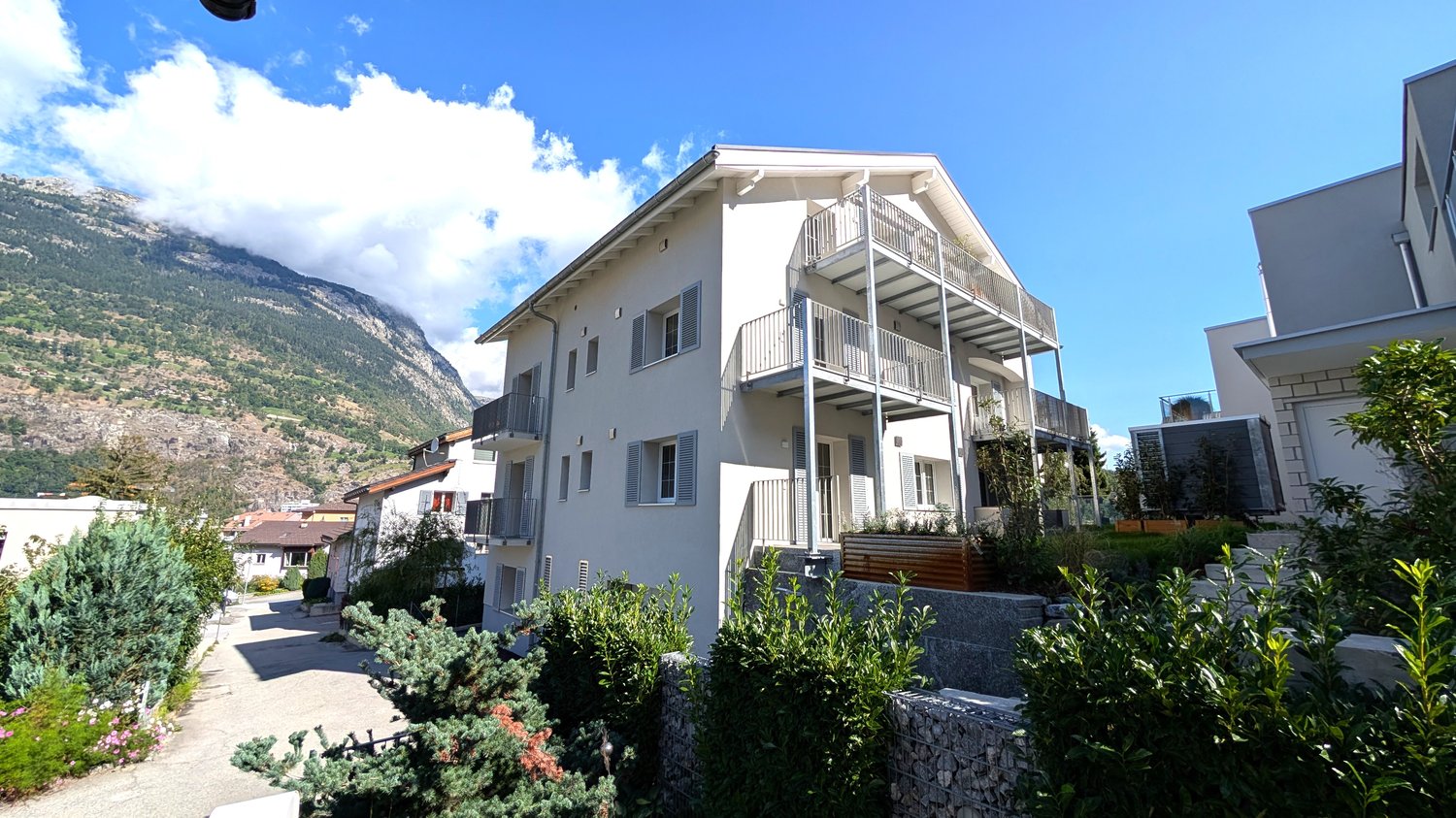 Three-story house with balconies on the upper floors, white exterior, modern architecture, surrounded by greenery and mountains