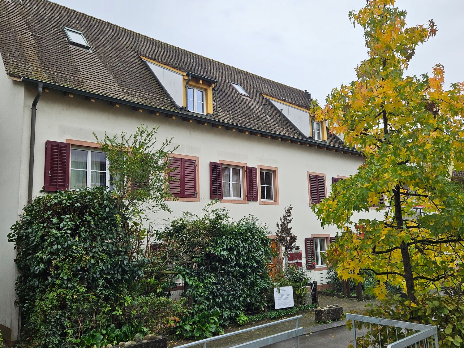 Raised ground floor white house with brown roof, multiple windows with red shutters, surrounded by green plants