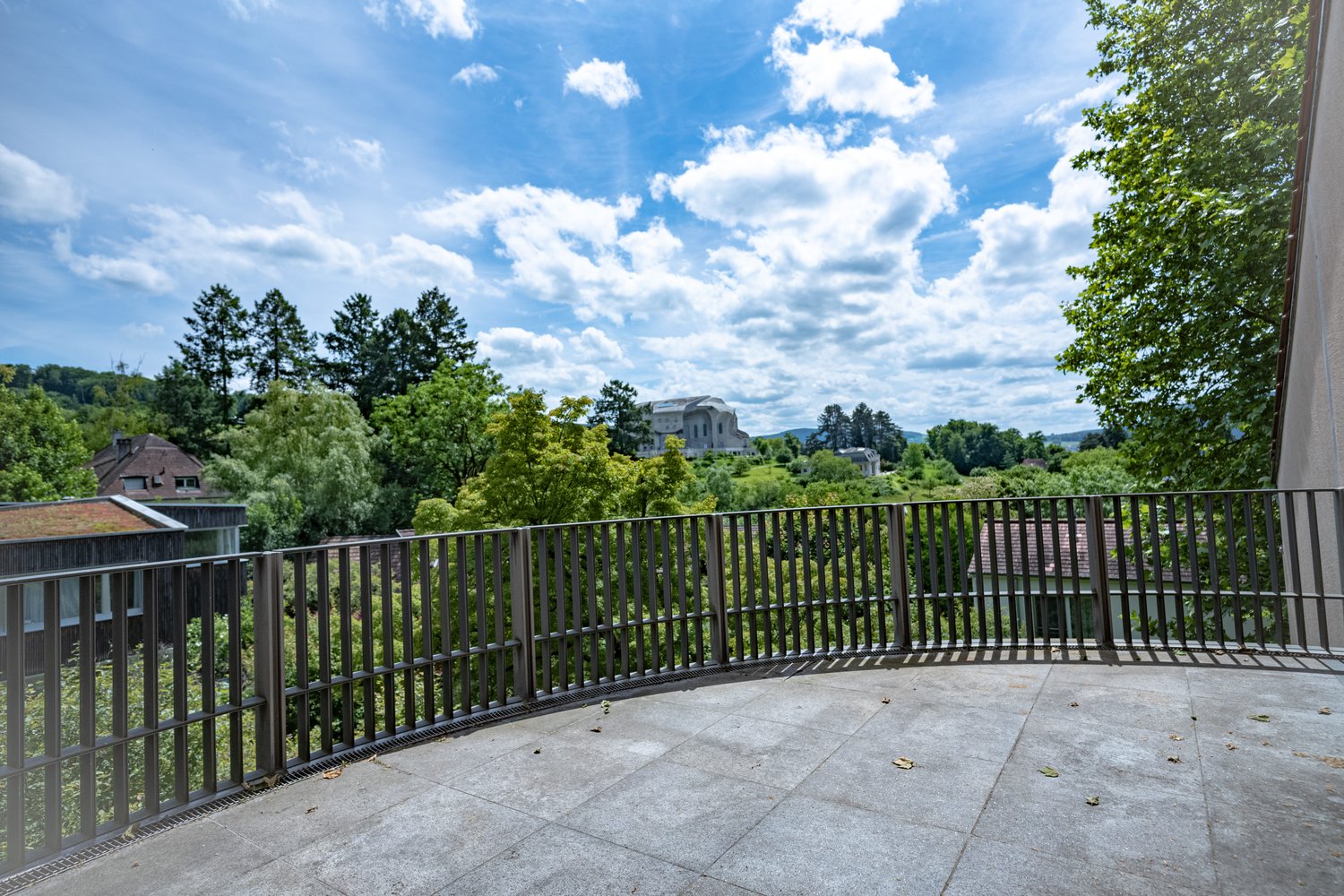 The image shows an outdoor terrace or balcony with a metal railing overlooking a scenic landscape. The terrace has a stone or concrete floor, and the view beyond the railing includes lush green trees, a building in the distance, and a partly cloudy blue s