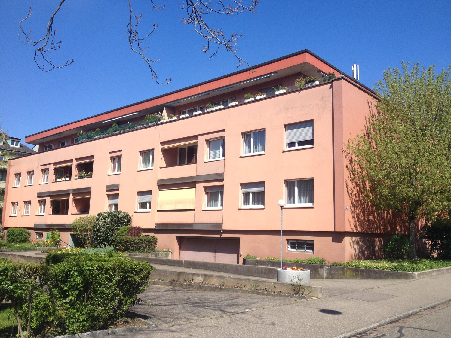 Apartment building, red facade, multiple windows, balconies, garage, garden, parking area