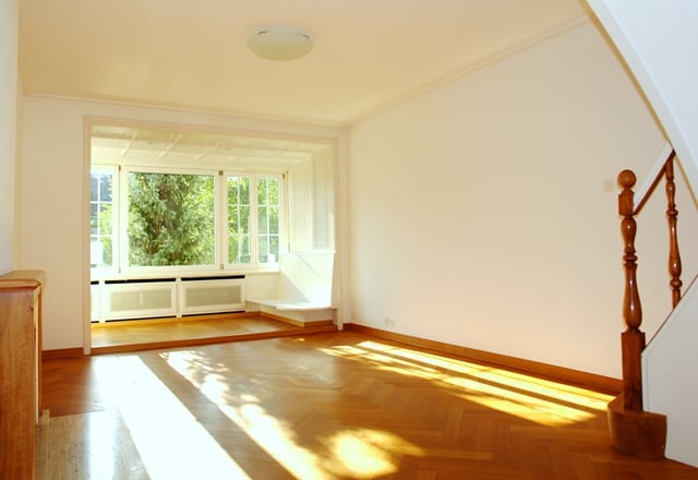 Empty room with wooden flooring, white walls, and a wooden staircase. There are large windows with a balcony and trees visible outside.