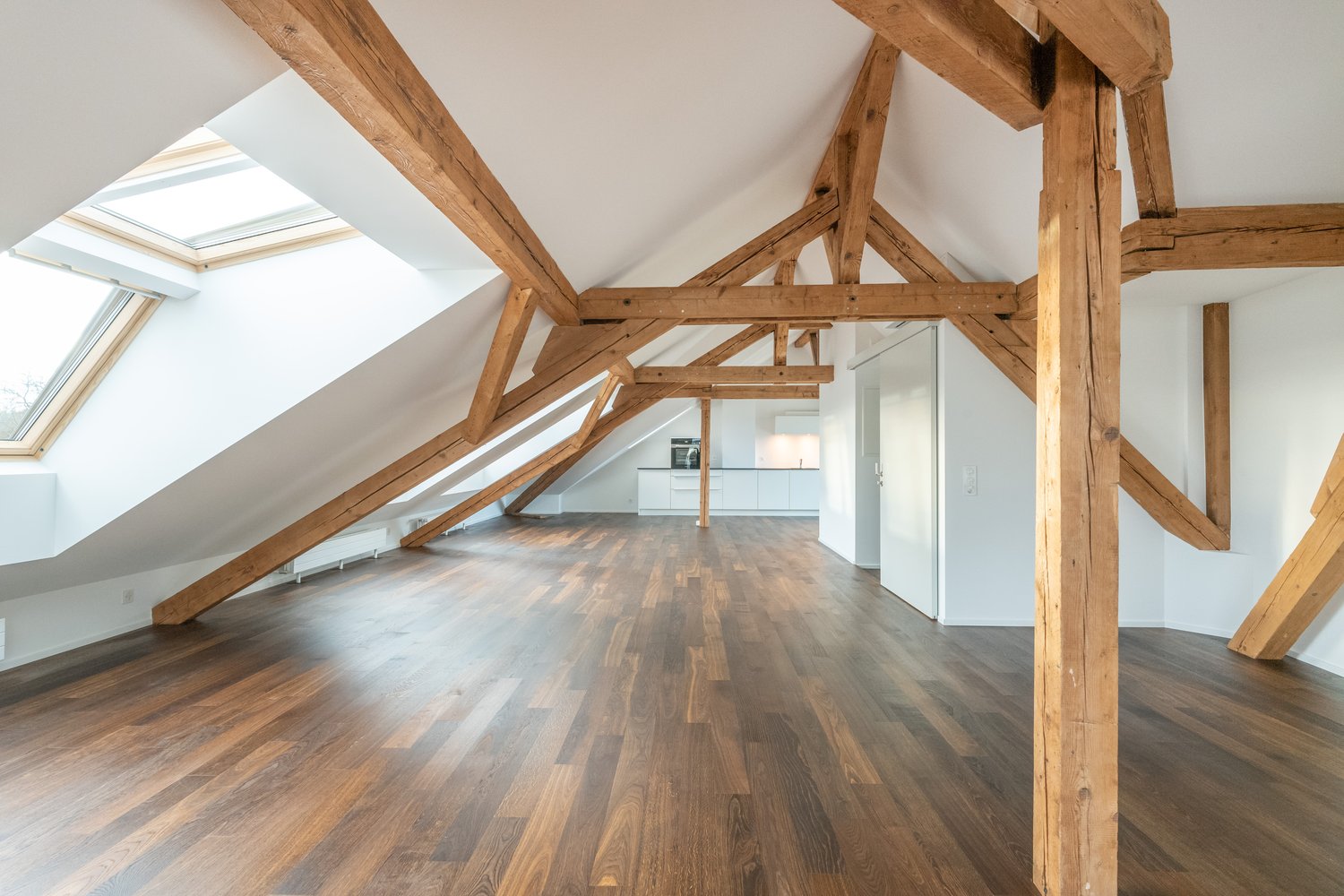Spacious attic room with exposed wooden beams, hardwood flooring, and a skylight providing natural light. The room appears to be an open-concept living space with a kitchen visible in the background.