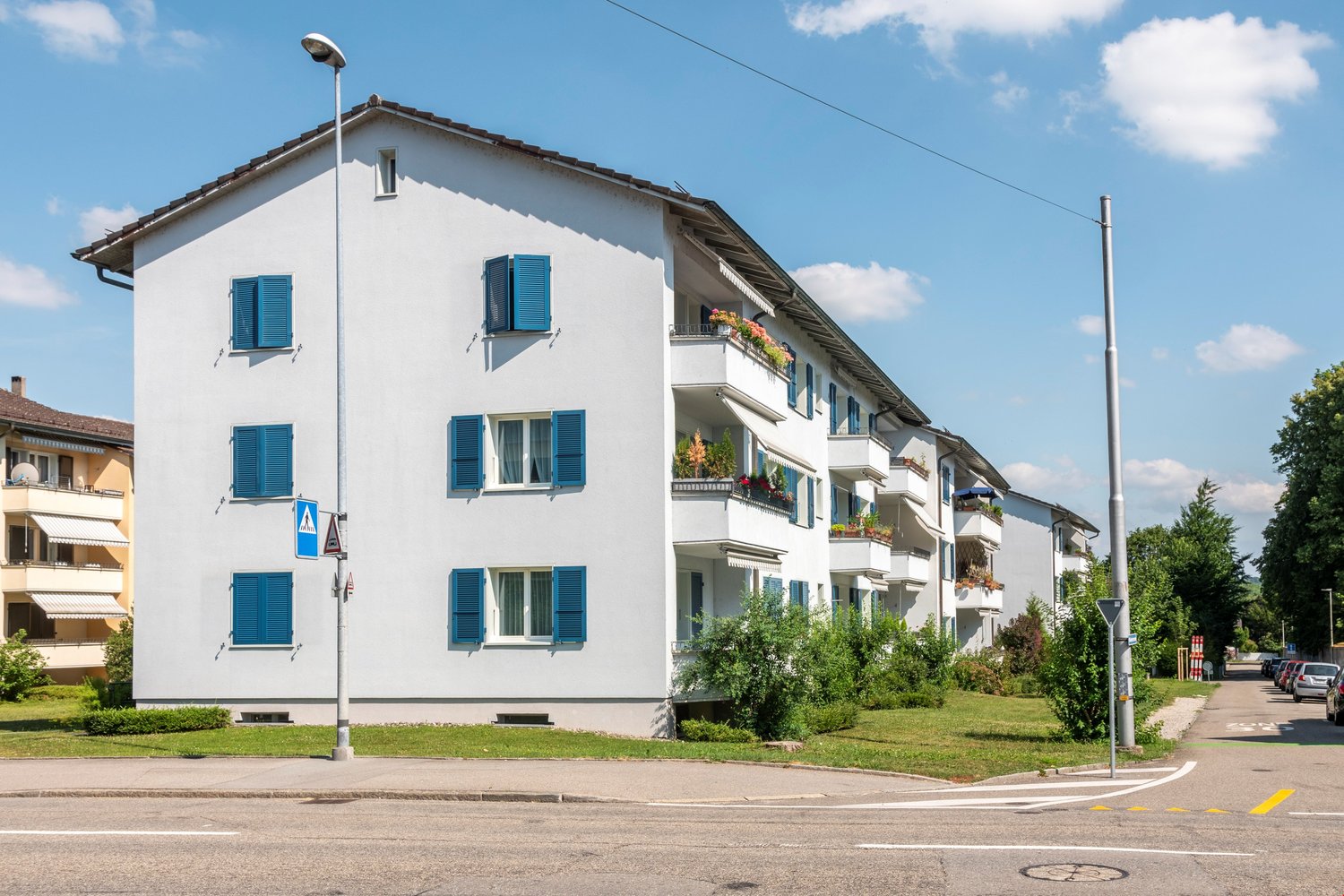 Multi-storey building, white exterior, blue shutters, balconies with plants, streetlight, street, parked cars.