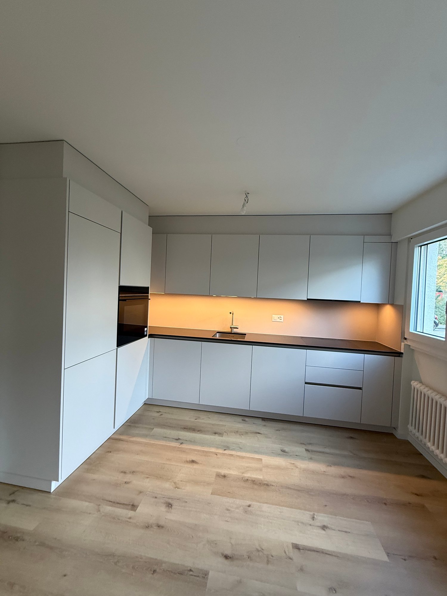 Modern white kitchen with wooden floors, integrated oven, built-in cabinets, modern lighting, and a window with a view
