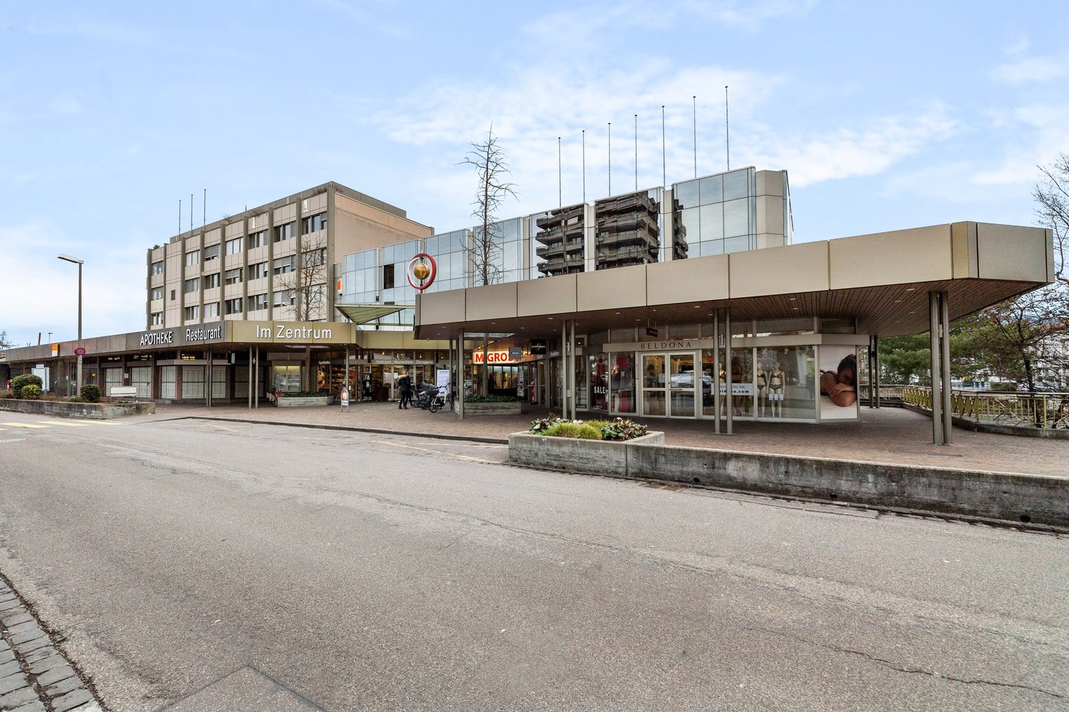 Commercial buildings with glass facades, name boards indicating 'Apoteke Restaurant', 'Im Zentrum', and 'Beldona', glass doors with signage, street light on the left, a few trees and plants in the foreground.