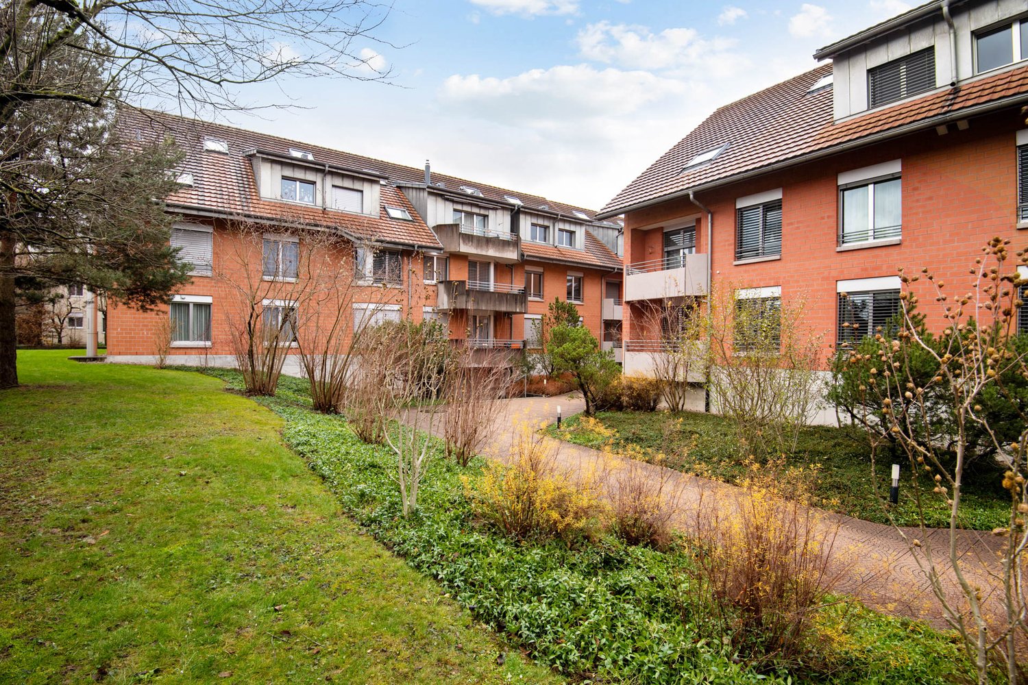A row of brick apartment buildings with balconies and windows, surrounded by a grassy area and shrubs.