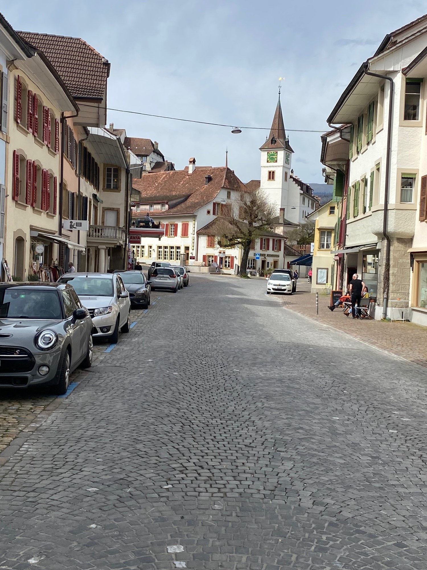 Cobblestone street, multiple parked cars, shops on both sides, a clock tower in the distance, a person sitting on a sidewalk, an umbrella hanging outside a shop.