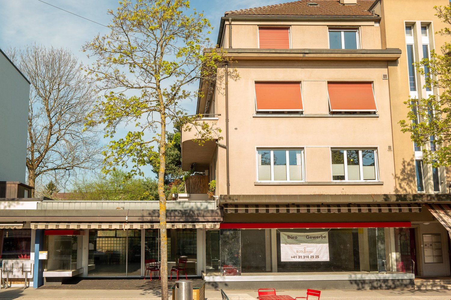 Three-story building, beige exterior, glass storefront, awnings, balcony on second floor, trees