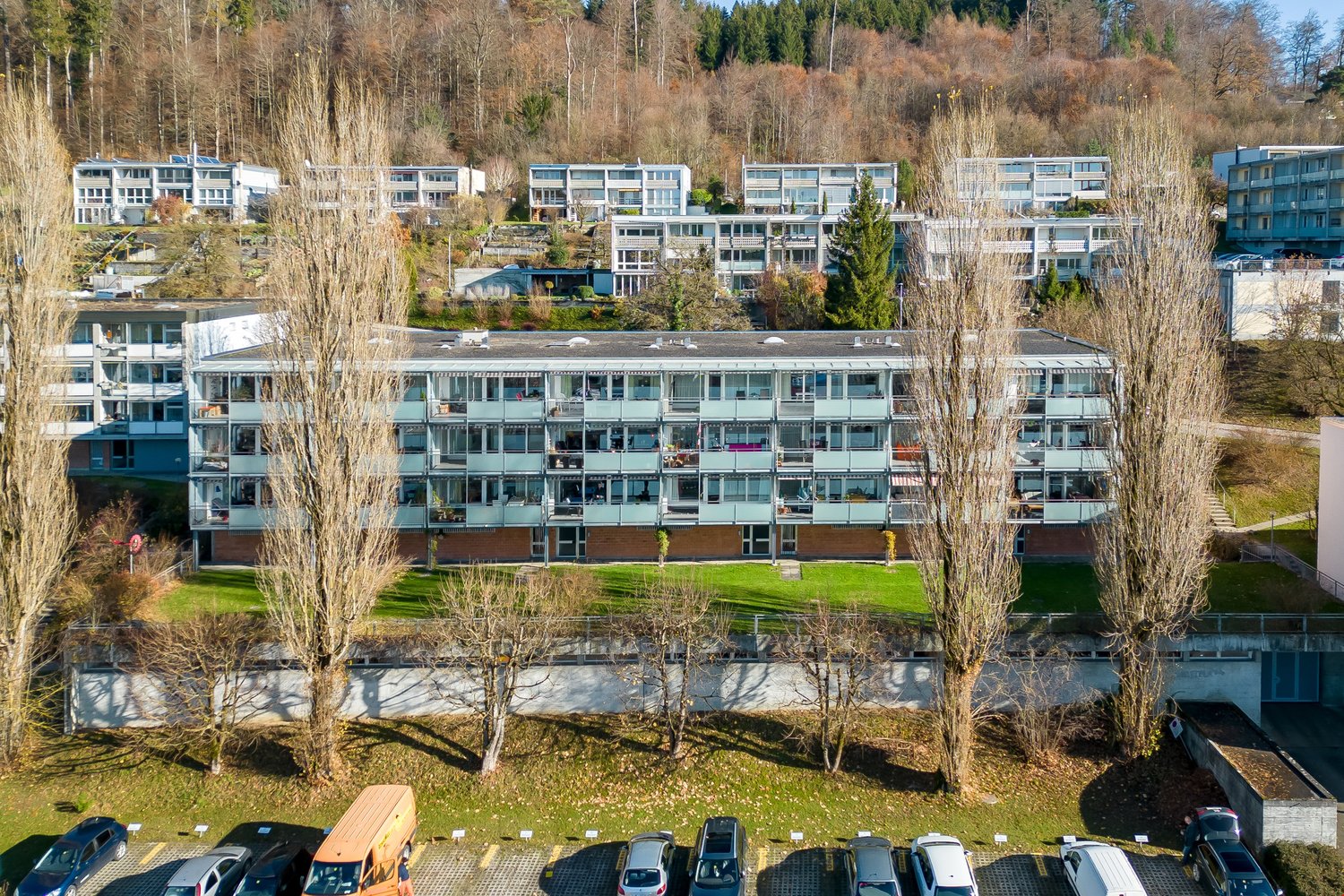 Apartment building with several floors, balconies, parking lot, green grass and many trees in the surroundings.