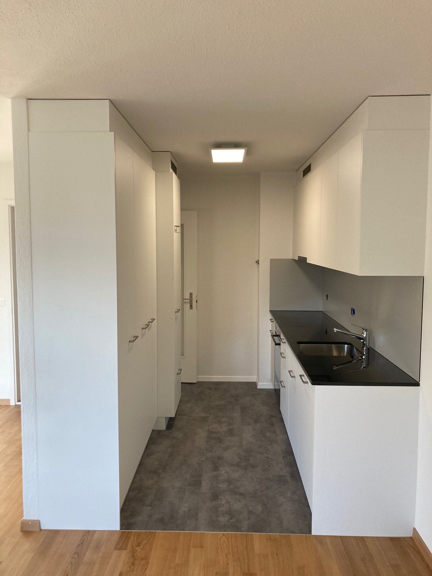 Modern kitchen with high-gloss white cabinets, a black granite countertop, a stainless steel sink, and a chrome faucet. Equipped with built-in appliances, ample storage space, and a sleek design. The kitchen is spacious and well-lit, with a ceiling lamp a