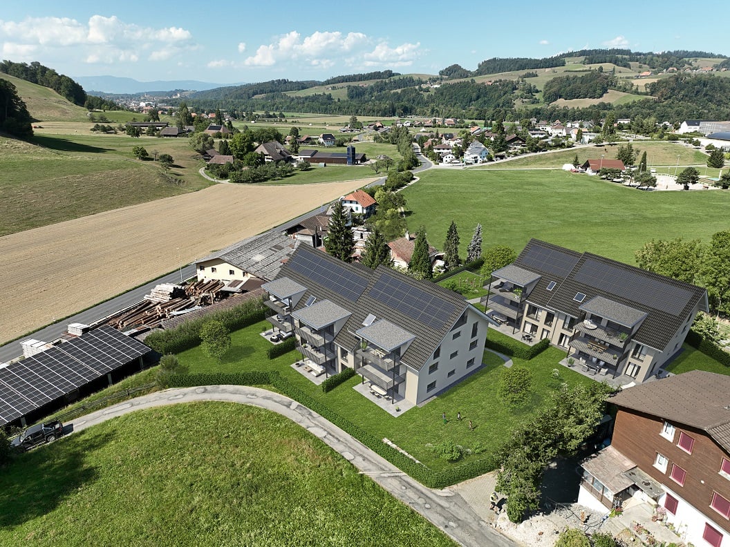 The image shows a small village or town nestled in a hilly, rural landscape. The buildings have solar panels installed on their roofs, indicating the use of renewable energy. The surrounding countryside is lush and green, with fields and forests visible i