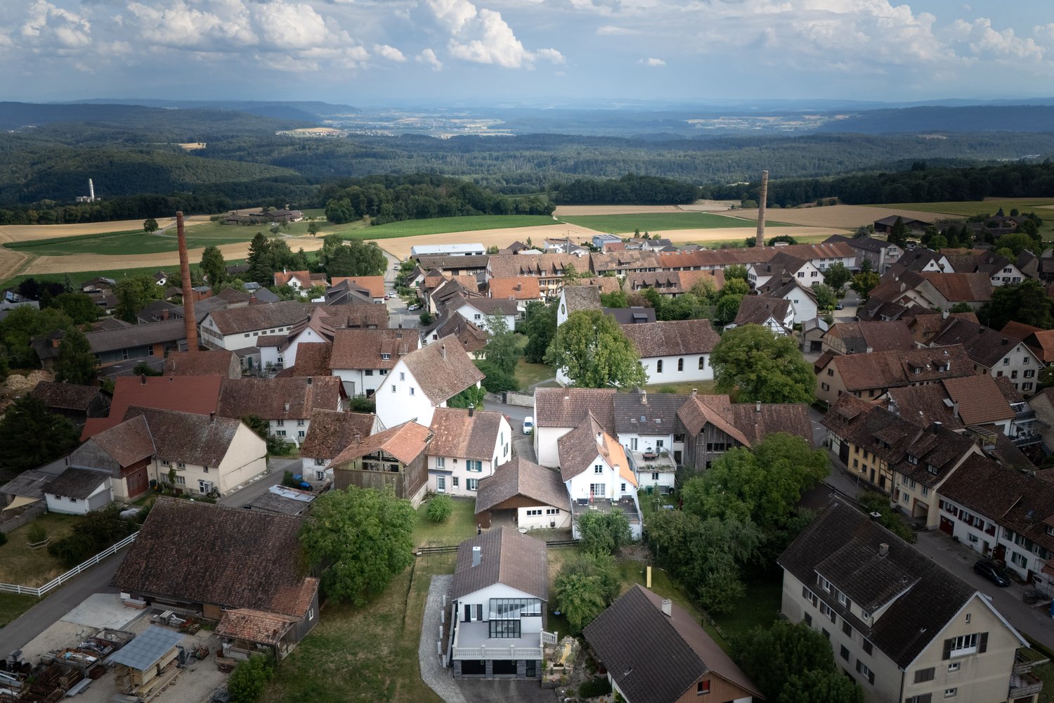 Aerial view of a picturesque German town nestled in a valley surrounded by rolling hills and forests. The town features traditional red-roofed buildings, narrow streets, and a church steeple. The landscape includes agricultural fields and a scenic rural s