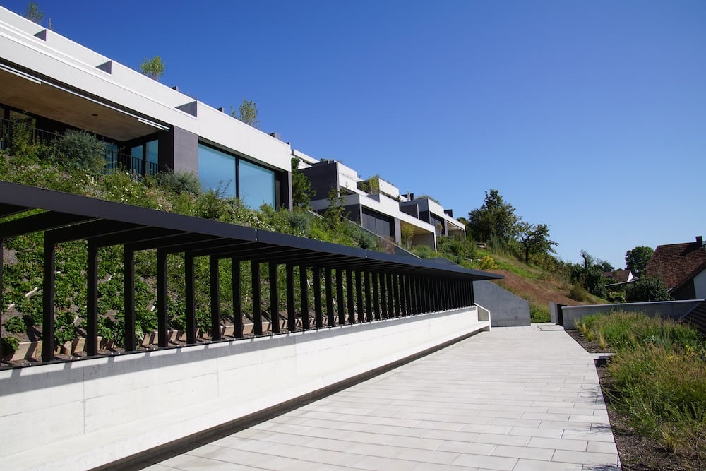 Modern multi-story building with balconies, surrounded by greenery and a paved walkway leading to the entrance.