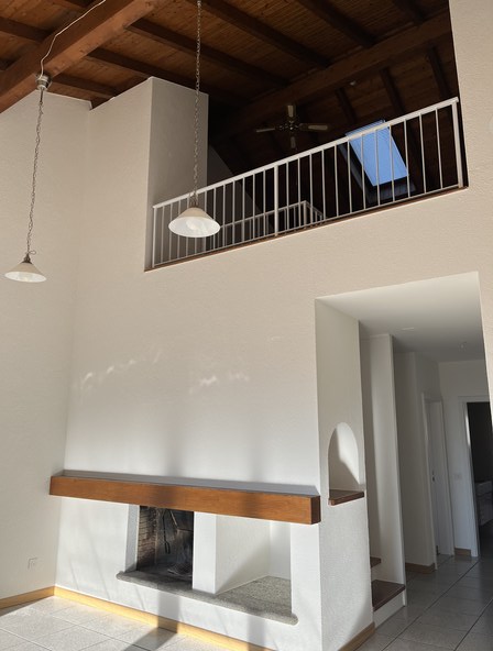 The image shows the interior of a room with a high ceiling and exposed wooden beams. There is a balcony or terrace visible through the railing on the upper level. The room has a minimalist design with white walls and a wooden bench or shelf below the balc