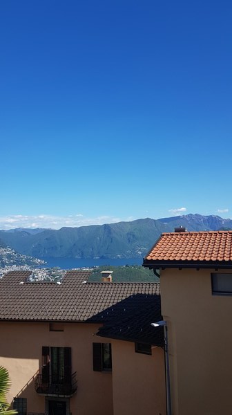 The image shows a scenic view of a mountainous landscape with a clear blue sky. In the foreground, there are rooftops of buildings with red tiles. The background features mountains and a body of water, likely a lake or a bay.