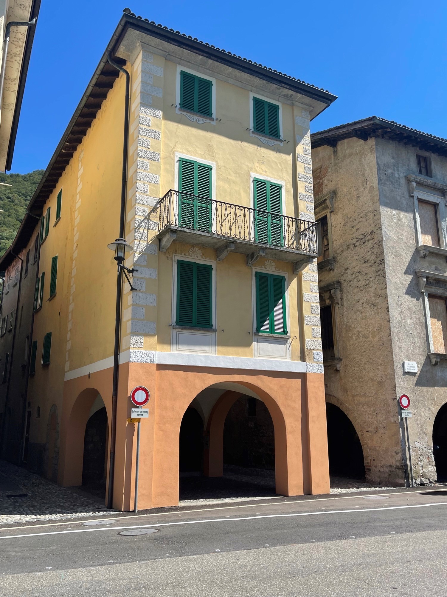 Two-story building with yellow exterior walls, green shutters, and a balcony on the second floor. The building has an arched entryway on the ground floor.