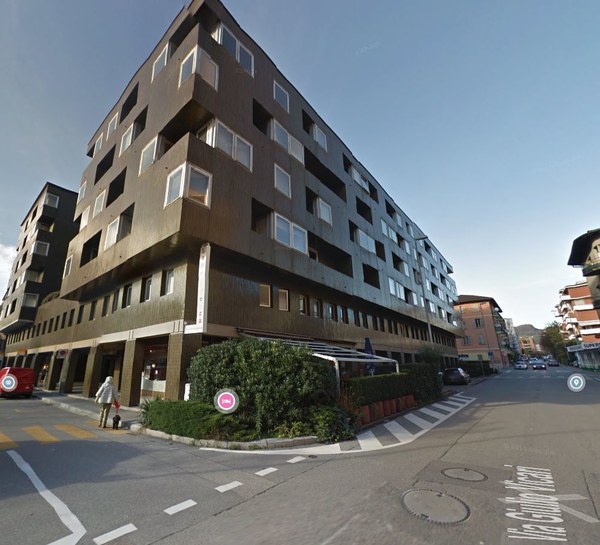 A multi-story residential building, with a modern architectural design and several windows. The ground floor features a retail space with signage and an awning. The building is situated on a street corner with greenery, and a crosswalk is marked.