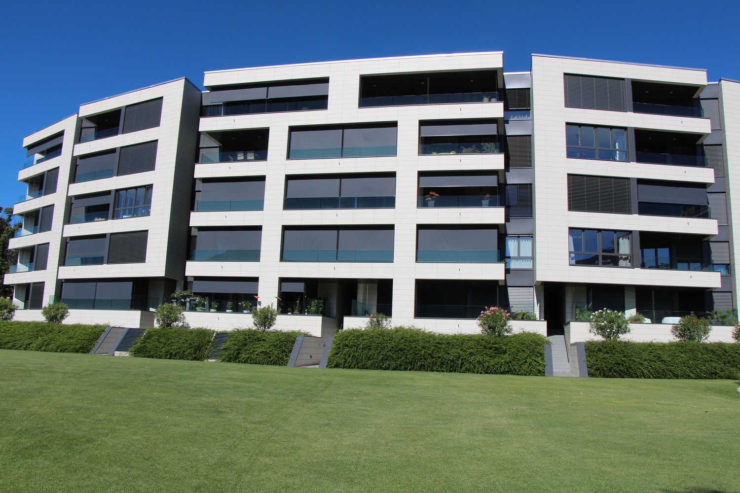 Multi-story apartment building, white exterior with glass balconies, greenery, modern design, large grassy lawn