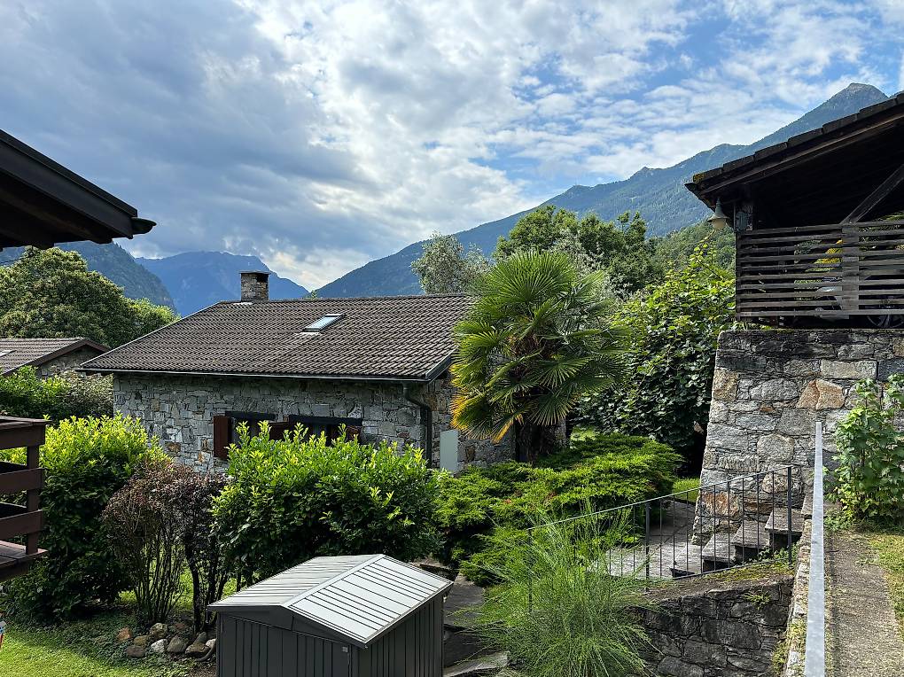 stone house with brown roof, stone walled exterior, solar panel on roof, surrounded by greenery, mountains in the background