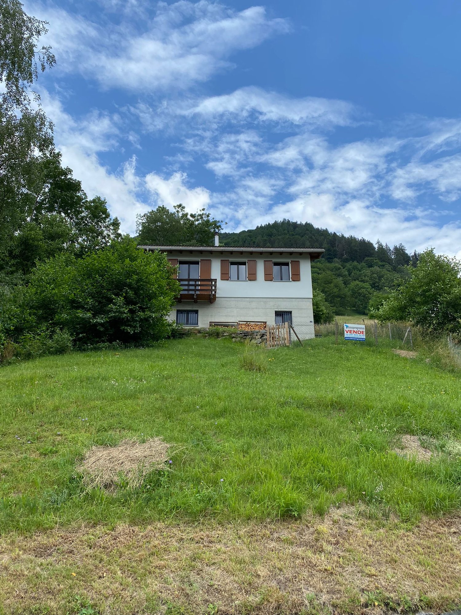 2-story house with a balcony, surrounded by green trees and a grassy field. There is a 'Vende' (For Sale) sign visible in the foreground.