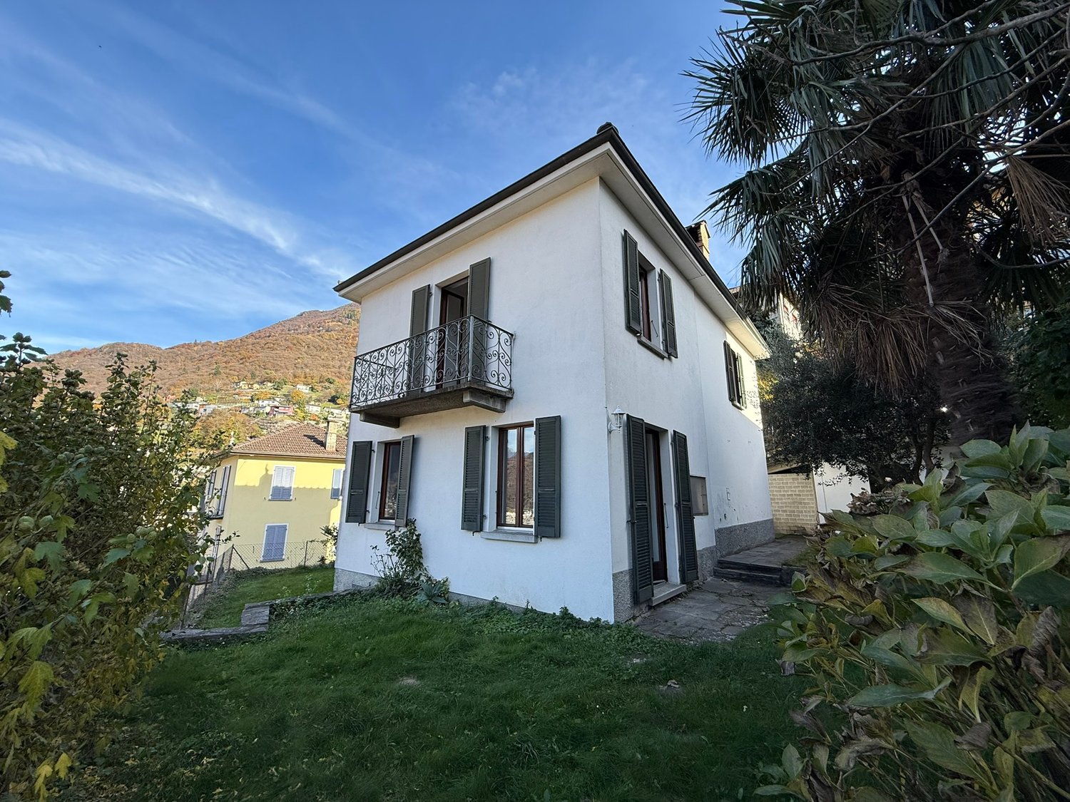 white house, 2 stories, balcony, green shutters, fenced garden, mountain in background