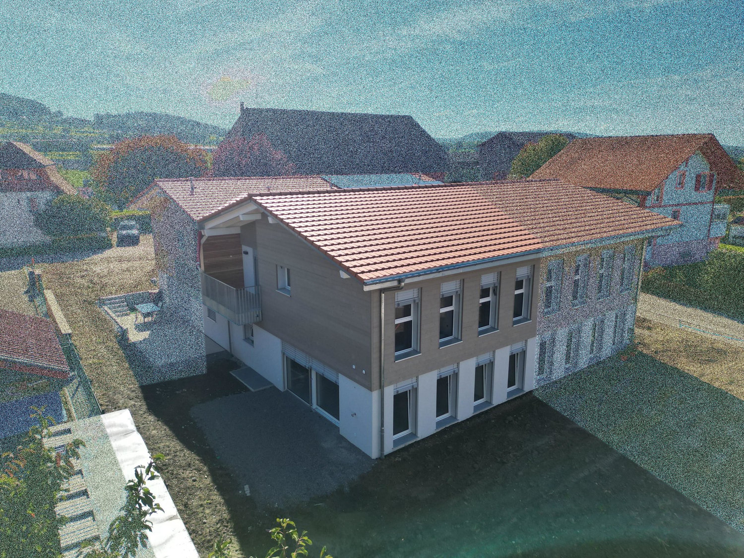 A modern two-story house with a tiled roof, large windows, and a balcony on the upper level. The house features a mix of stone and wood exterior finishes and is situated in a residential area with neighboring houses visible in the background.