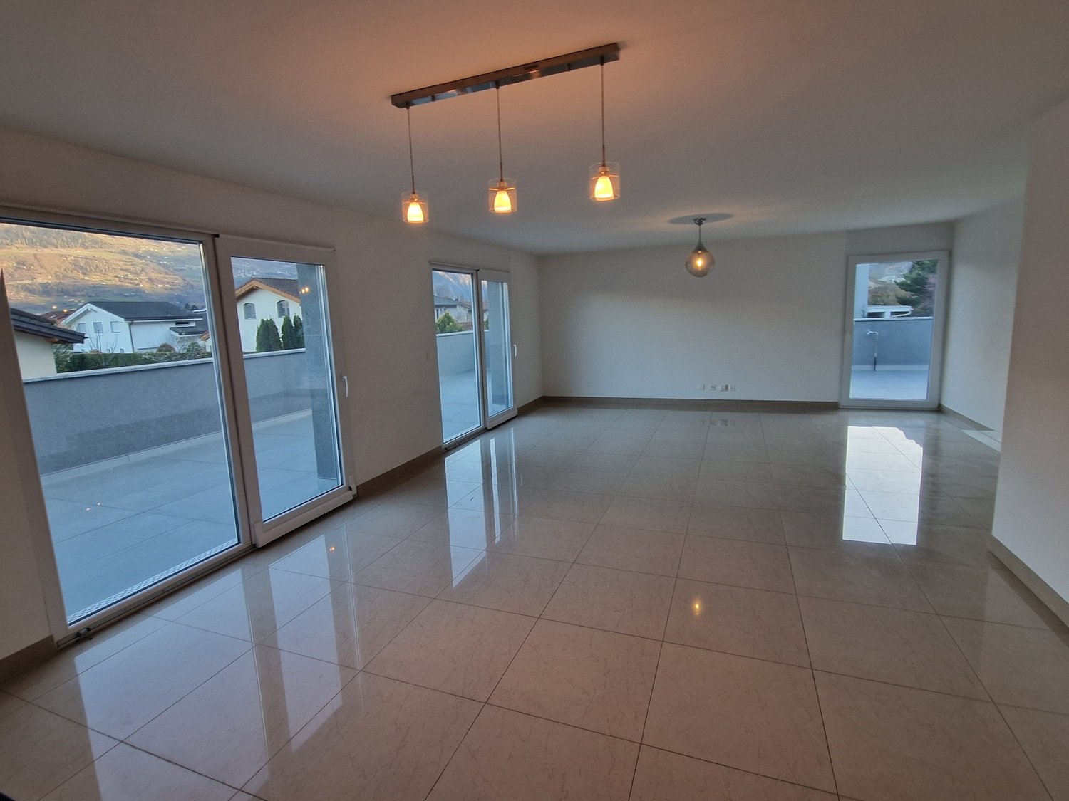 Spacious open-plan living area with large windows, hardwood floors, and modern pendant lighting fixtures. The room has a clean, minimalist design with a neutral color palette.