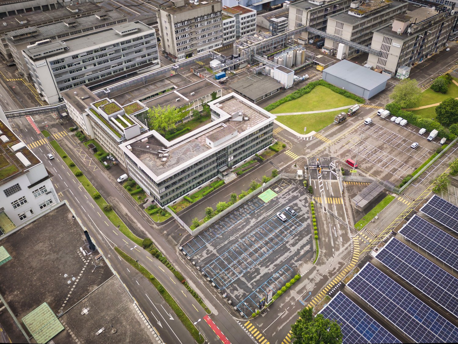 Industrial complex with multiple buildings, parking lot, solar panels, greenery, rooftop garden, wide roads, street lights