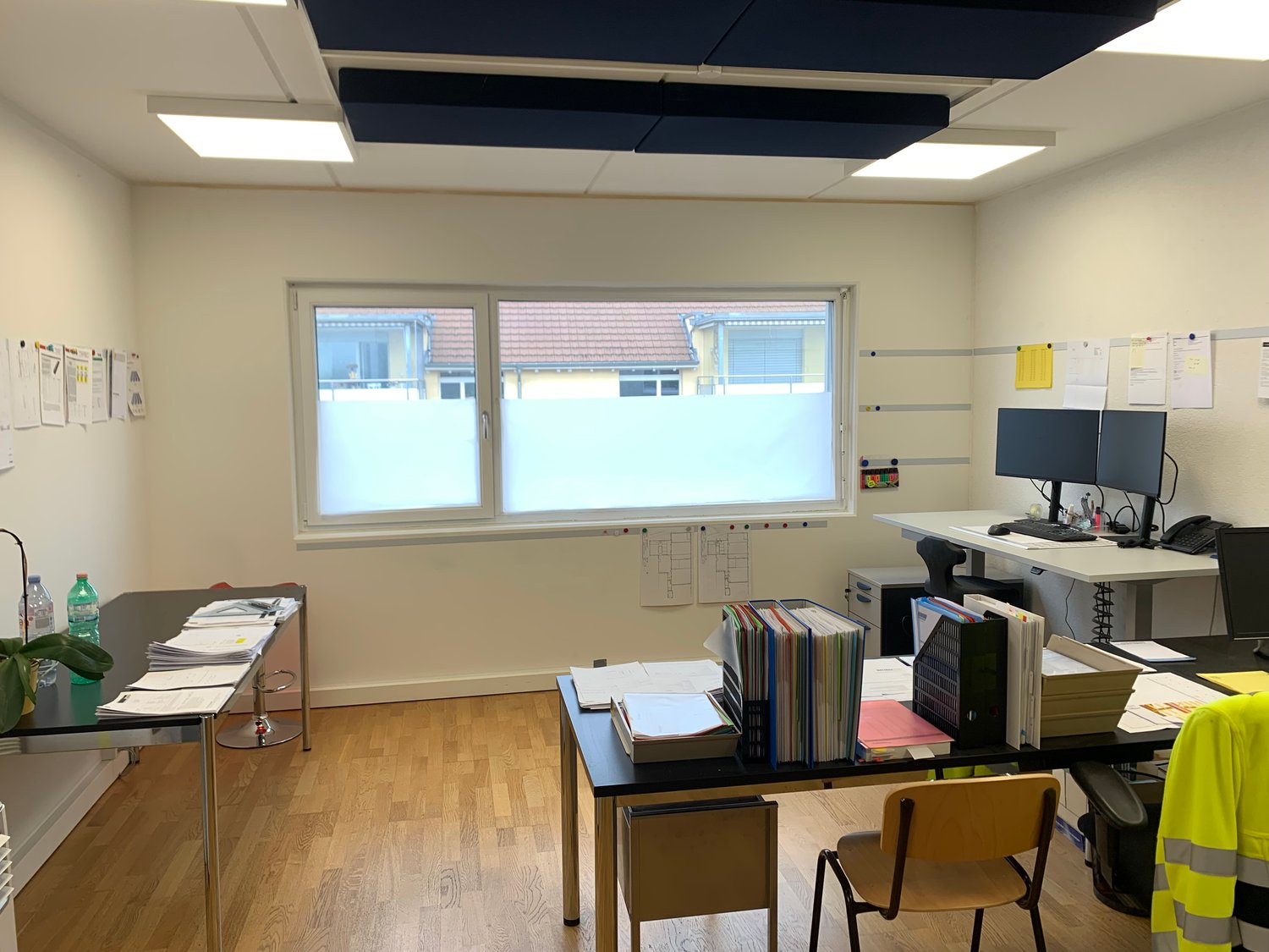 A modern office room with a desk, shelves, a chair, and a window. The room has a clean and organized appearance with white walls and wooden flooring. The desk contains monitors, keyboards, telephones, and various office supplies. There are two tables with