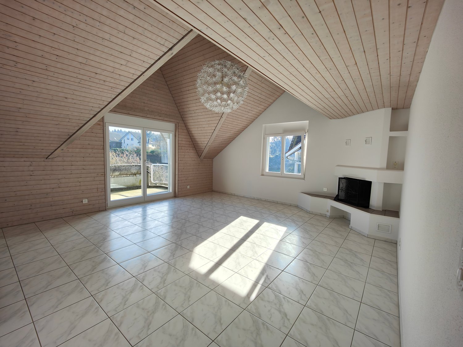 Spacious attic living room with vaulted wooden ceiling, large windows, and sliding glass door leading to a balcony or terrace. The room has a tiled floor and a modern light fixture hanging from the ceiling.