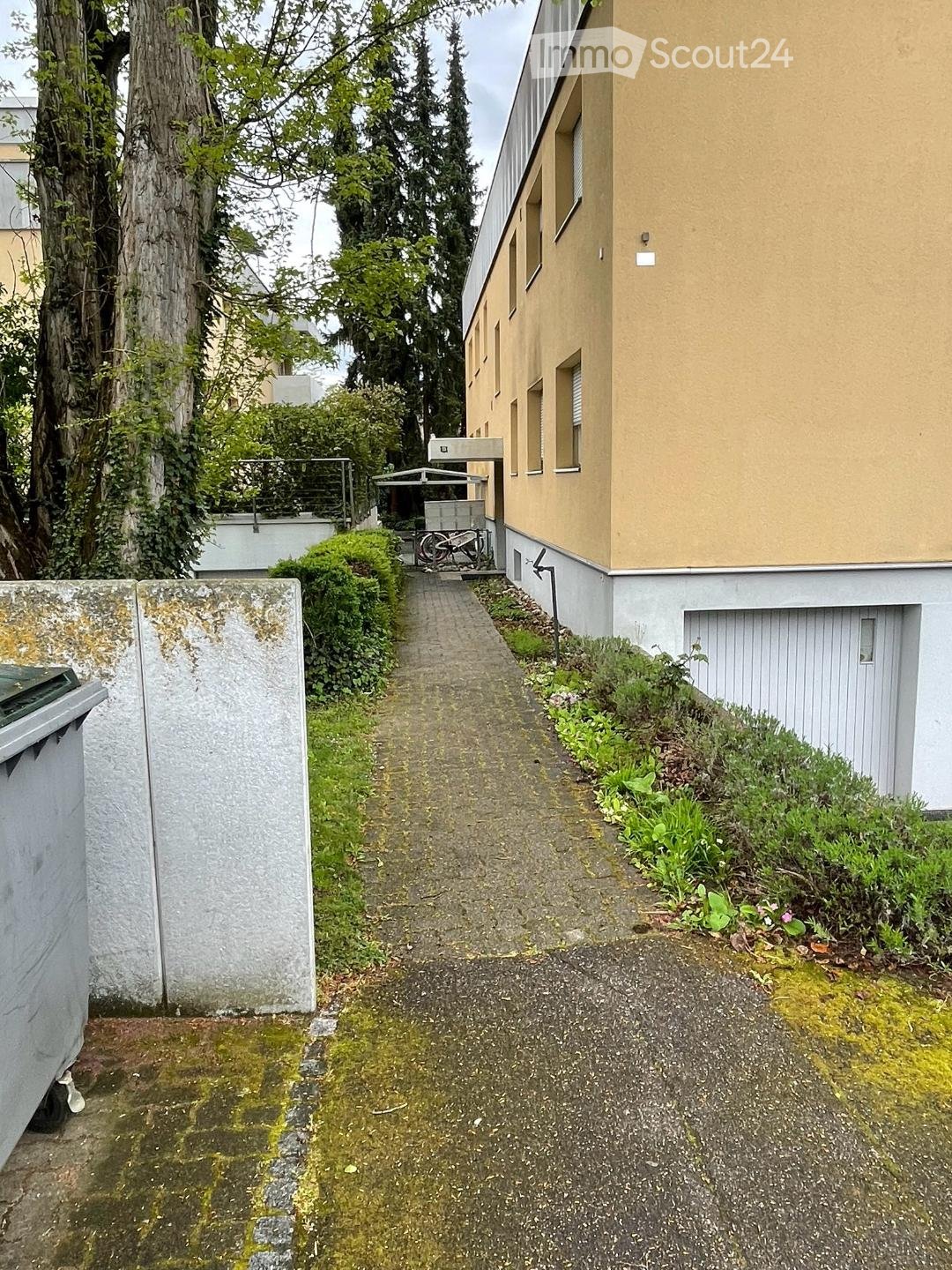 A multi-story building with a yellow exterior, surrounded by trees and greenery. There is a paved walkway leading to the building, with some vegetation along the sides.