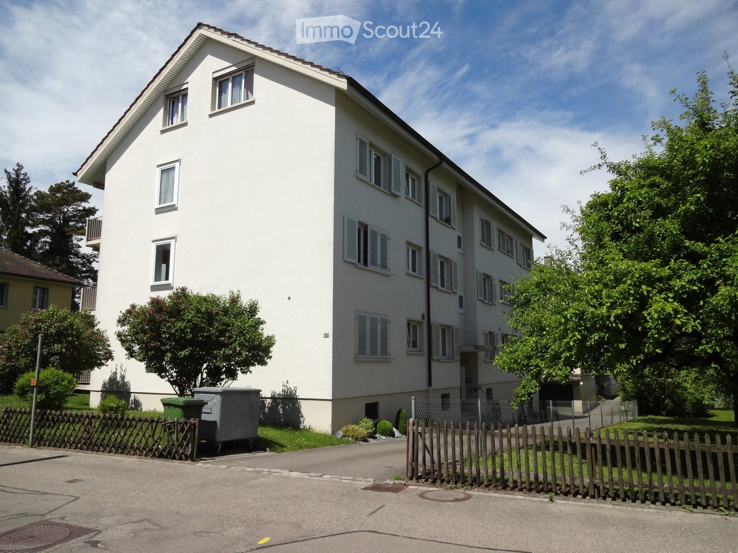 white multistory building, multiple windows, red roof, wooden fence, shrubs and trees
