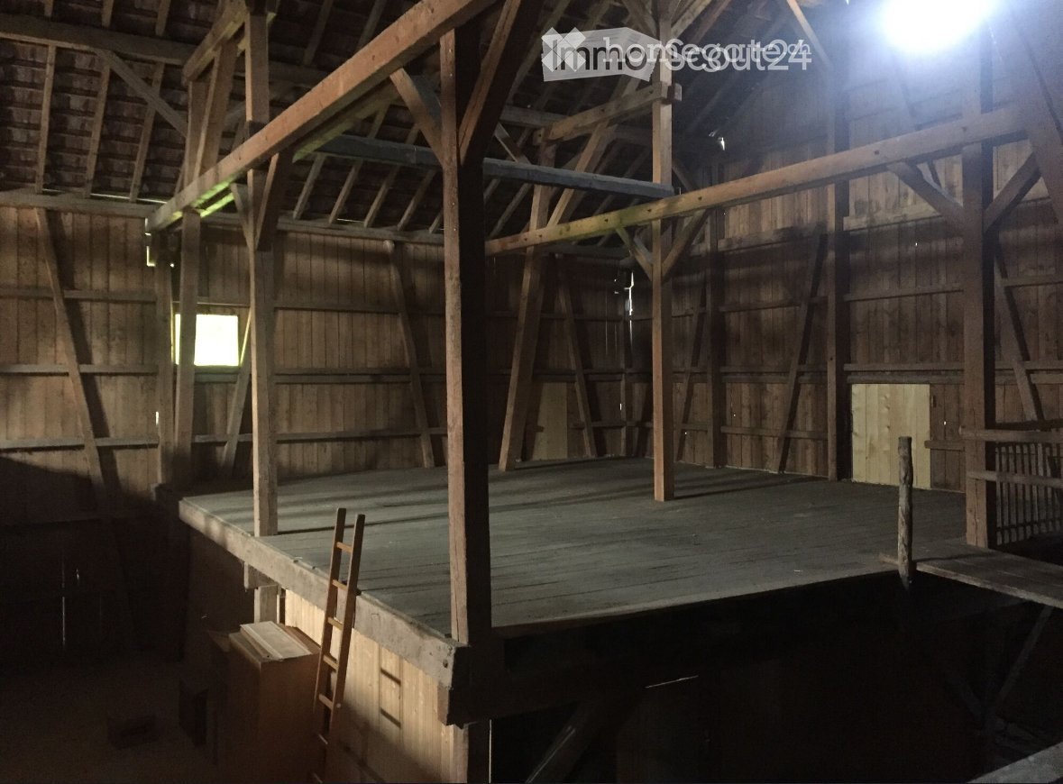 The image shows the interior of an old, wooden barn. The structure is made of wooden beams and supports, with a high ceiling and a loft area accessible by a ladder. The floor appears to be made of wooden planks. The overall atmosphere is dark and rustic,
