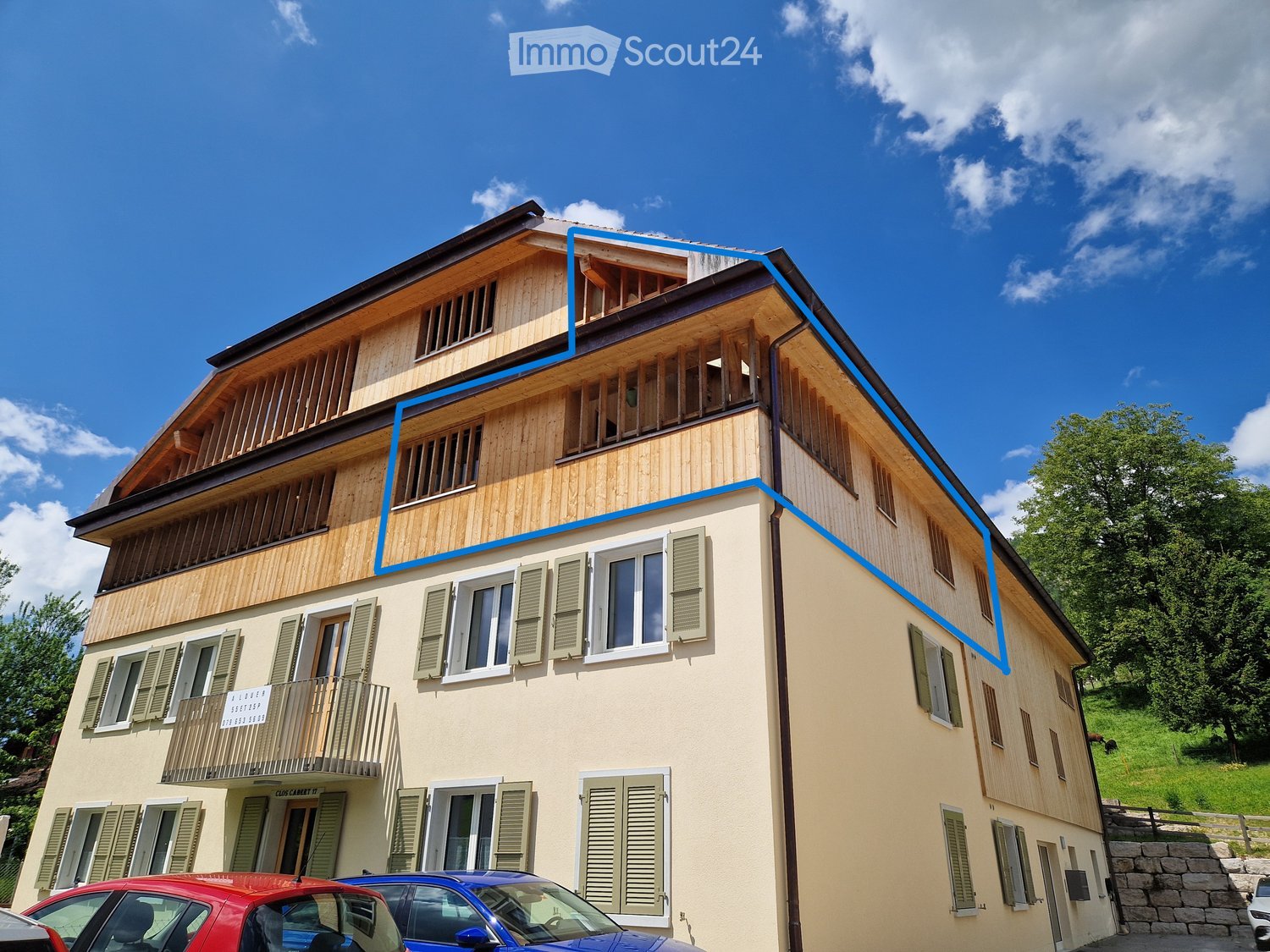 3-story wooden building with balconies, beige exterior walls, blue trim, and a sloped roof. There are several windows and a parking area in front of the building.