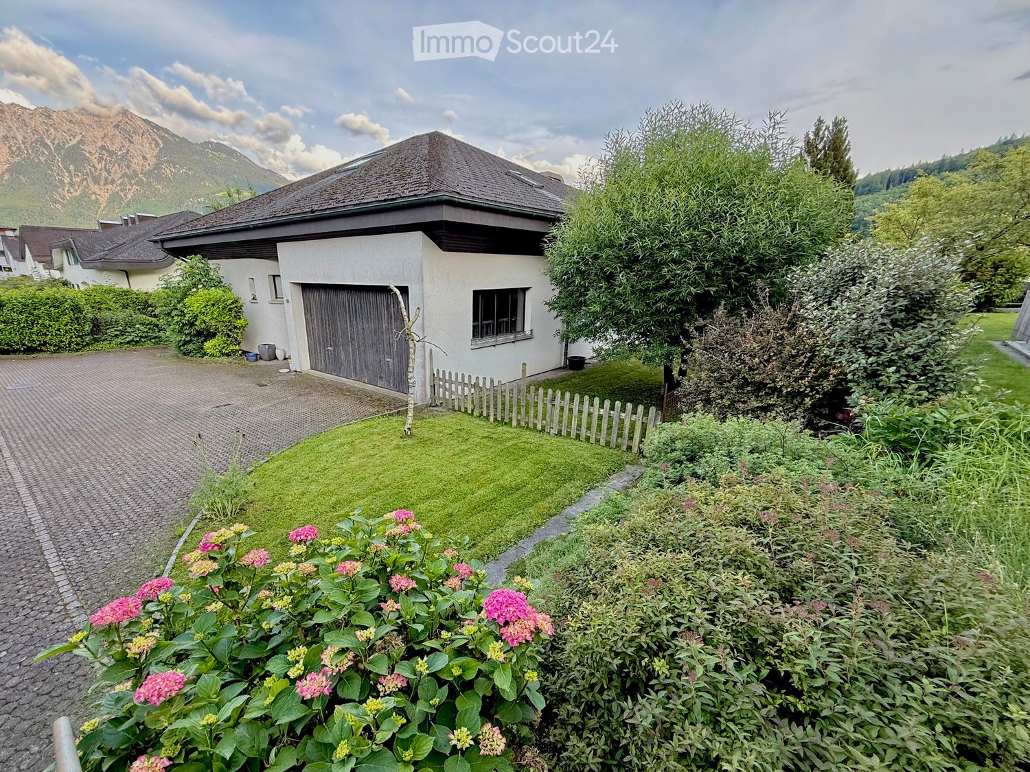 Single-story house with a gabled roof, surrounded by a well-maintained garden with lush greenery, flowering plants, and a paved driveway leading to a garage.